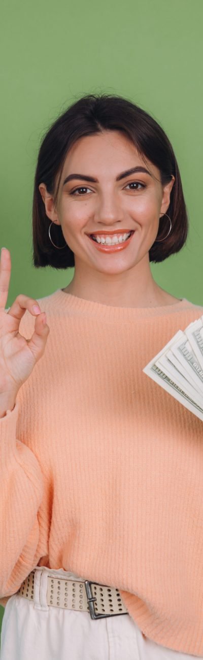 Young woman in casual peach sweater  isolated on green olive background  lucky holding fan of 100 dollar bills being excited to win cash prize showing OK sign